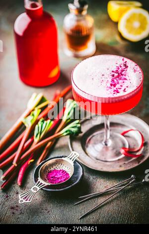 Beautiful pink cocktail with frothy top in coupe glass with sprinkled berry powder garnish, fresh rhubarb stalks and syrup in background Stock Photo