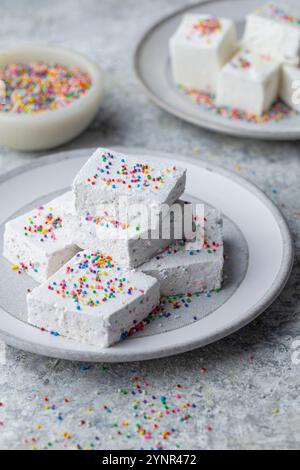 Side shot of a stack of square white marshmallows topped with colorful rainbow sprinkles on a pale gray plate. A soft focus bowl of sprinkles and more Stock Photo