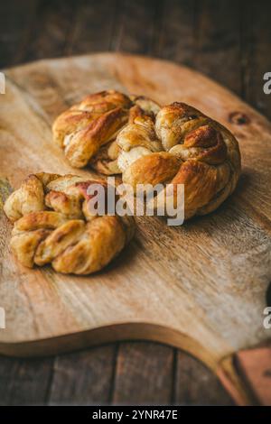 Three freshly baked, golden brown Cardamom Buns on a wooden cutting board. The background is a dark brown wooden table, giving the image a warm and in Stock Photo