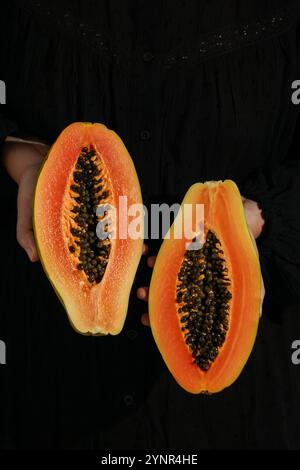 fresh, ripe papaya sliced ​​open in woman's hands, on black background, harsh sunlight Stock Photo