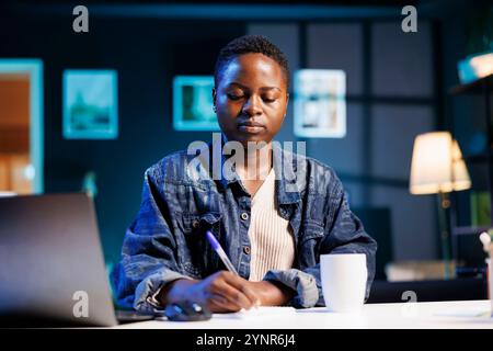 Young black woman studying while using her laptop and taking notes on a notepad. African American female entrepreneur browsing the internet and writing down new business ideas. Stock Photo