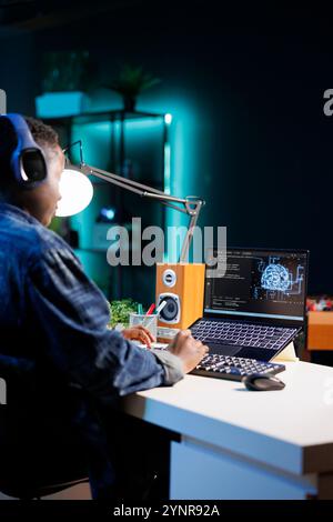 Female developer using headphones while coding on a laptop with a blue interface showing a neural network. Black woman manages cyber security and monitors the network in a futuristic data center. Stock Photo
