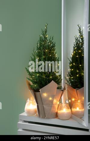 Small spruce tree decorated with Christmas lights and burning candles on white radiator cabinet near green wall Stock Photo