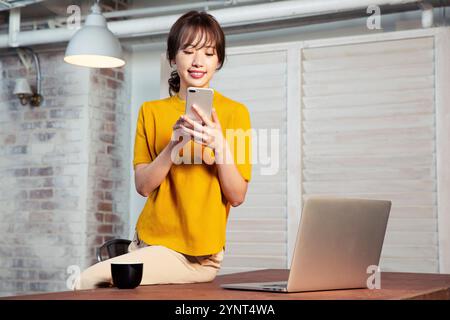 Woman in her 20s sitting at a desk operating a smartphone Stock Photo
