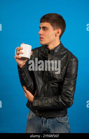 Young man wearing black leather jacket drinking from white mug on blue background Stock Photo