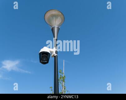 Security video camera mounted on a lamppost outdoors against a blue, partly cloudy sky. Stock Photo