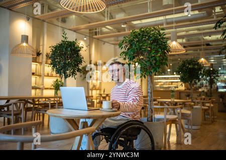 Interested man with disability drinking coffee in cafe freelance working online, watching on laptop. Stock Photo