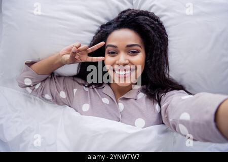 Young woman in polka dot pajamas making peace sign while lying in bed, enjoying morning leisure time Stock Photo