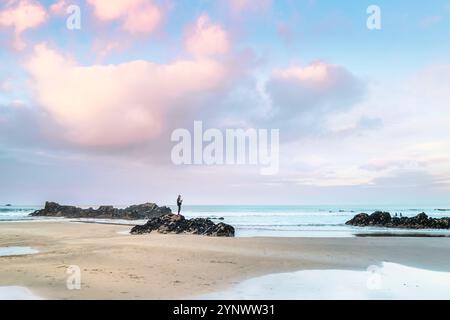 Pastel coloured sky over the figure of a person standing on rocks exposed by a low tide on Gt Western Beach on the coast of Newquay in Cornwall in the Stock Photo