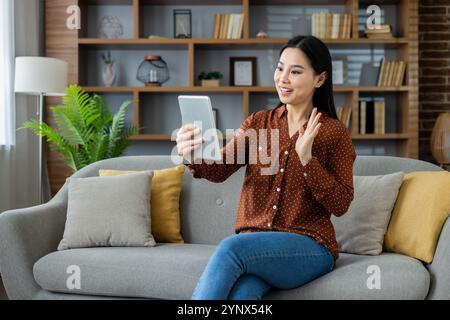 Asian woman smiling, sitting on sofa using tablet for virtual conversation. Waving in greeting, enjoying relaxed comfort at home. Highlighting virtual communication, technology, connection. Stock Photo
