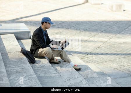Side view of Asian male IT developer typing codes on laptop working outside in sunlight while sitting on concrete steps against paved square in city s Stock Photo