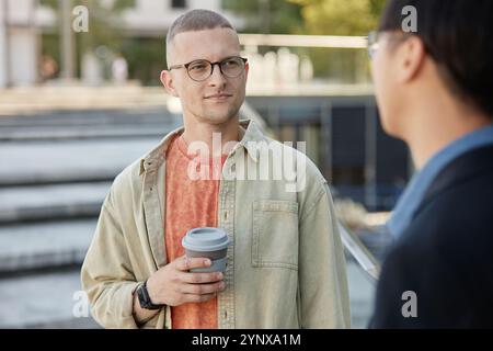 Medium shot of casually dressed man with reusable cup in hand meeting with friend or colleague while enjoying communication and nice day outside in ci Stock Photo