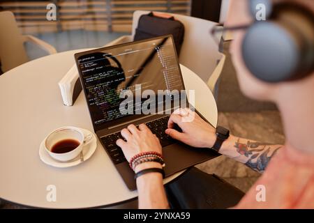 Over shoulder shot of male software developer writing code on laptop sitting at cafe table with cup of coffee at hand during coffee break or lunch, co Stock Photo