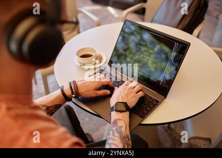 Over shoulder shot of male IT developer writing code on laptop sitting at cafe table with cup of coffee at hand during coffee break or lunch, copy spa Stock Photo