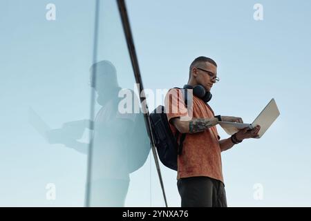 Side view shot of concentrated adult male IT programmer with tattoo on arm wearing backpack typing on laptop while holding computer with one hand agai Stock Photo