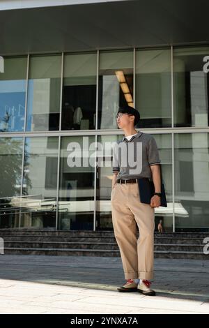 Vertical shot of fashionable Asian male IT programmer in office clothing holding laptop standing in sunlight next to business building while posing wi Stock Photo