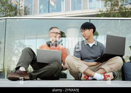 Candid shot of two smiling IT programmers from different ethnic backgrounds having fun while working on laptops collaboratively sitting on pavement ou Stock Photo