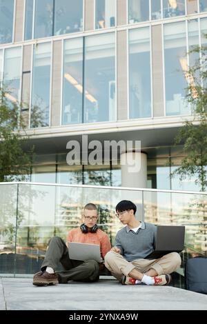 Vertical shot of diverse team of two male IT programmers working on laptops collaboratively sitting on pavement outside in city street against busines Stock Photo