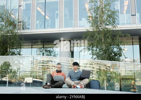 Wide angle shot of two male IT programmers from different ethnic backgrounds working on laptops collaboratively sitting on pavement outside in city st Stock Photo