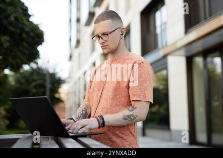 Medium shot of concentrated male IT programmer in wired headphones using laptop typing on keyboard while working at stand up desk in city street on bl Stock Photo