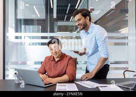 Team working on business project in office. Colleagues using laptop and smartphone for collaborative tasks, reviewing documents, engaging with technology for efficient workflow. Stock Photo