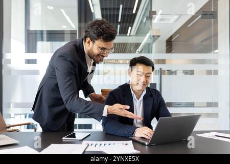 Businessmen collaborating in office. Focused team discussing project at laptop. One standing, one sitting, with smartphone and documents on table. Professional teamwork concept. Stock Photo