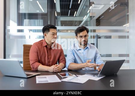 Business professionals in meeting discussing project. Colleagues collaborating over laptop, emphasizing teamwork, technology, strategy. Modern office environment enhances professional interaction. Stock Photo