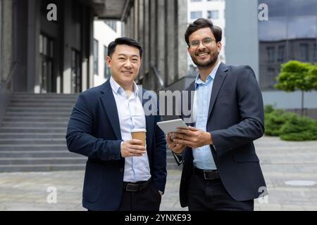 Two businessmen having an outdoor meeting, one holding coffee, other with digital tablet. Both smiling in business attire, representing teamwork, cooperation, and professional interaction. Stock Photo