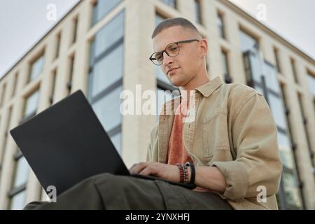 Low angle shot of handsome male software engineer wearing glasses writing code developing applications on laptop while sitting outside in front of bus Stock Photo