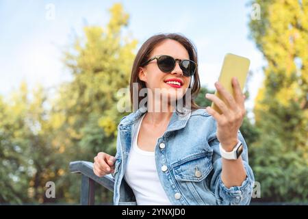 Urban woman in a denim jacket and sunglasses using a smartphone outdoors, with blurred greenery in the background. Stock Photo