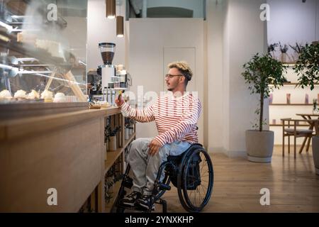 Concentrated disabled man in wheelchair pays for purchase in coffee shop with terminal, credit card Stock Photo