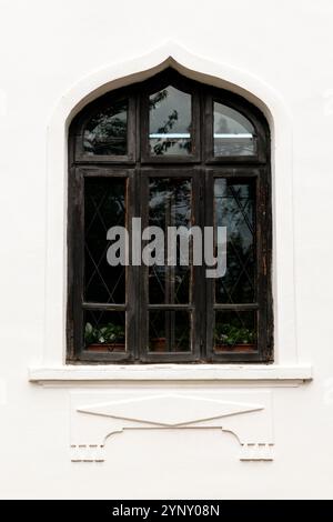 Elegant black arched window with intricate detailing on a white textured wall, reflecting surrounding greenery and sky, showcasing a timeless architec Stock Photo