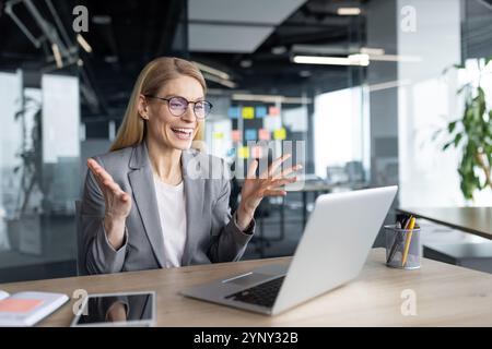 Mature woman businesswoman excitedly interacts during online meeting through laptop in modern office. Engaged in video call with enthusiasm, utilizing technology for remote work collaboration. Stock Photo