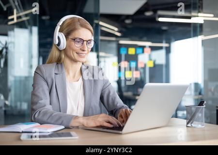Mature woman businesswoman working online wearing headphones, focused on laptop in modern office. Engaged in virtual meeting or remote work setup, portraying professional environment and technology. Stock Photo