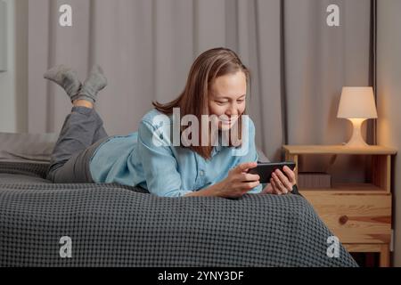 A smiling young woman lying down on bed and using smart phone chatting in social media, watching video content, texting messages, playing games, using Stock Photo