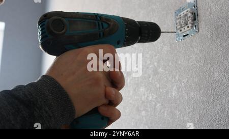 male hands holding cordless screwdriver and fixing structure for electrical switch or socket on wall with gray wallpaper Stock Photo