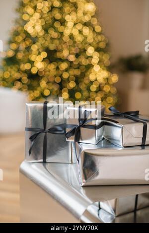 Close-up of a stack of Christmas gifts wrapped in silver paper in front of an illuminated Christmas tree Stock Photo