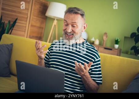 Senior man enjoying a relaxing day at home, engaging in a cheerful video call with friends on his laptop Stock Photo
