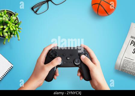 Top view flat lay composition of a player holding a gamepad on a desk with a ball, newspaper, glasses, plant, and notepad, perfect for creative gaming Stock Photo
