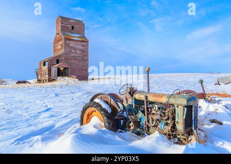 An old tractor is sitting in the snow next to a grain silo. The tractor is rusted and old, and the silo is also old and abandoned. The scene is quiet Stock Photo