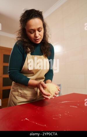 A woman in an apron skillfully kneads dough on a red table in a warm kitchen environment. The image captures a sense of focus, dedication, and the art Stock Photo