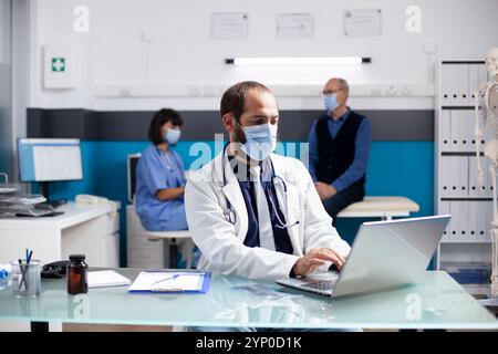 Male general practitioner sits in hospital office, using laptop and planning treatments for his patients during appointments. Doctor checking emails and preparing medical consultations in clinic. Stock Photo