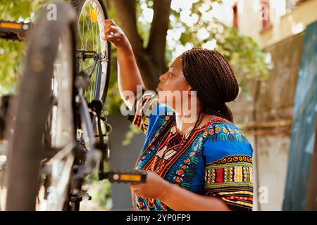 Sports-loving african american woman holding professional equipment for bike maintenance in home yard. Determined young female cyclist focuses on repairing bicycle wheel with specialized spanner. Stock Photo