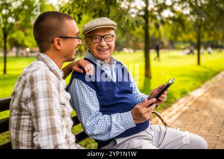 Portrait of happy grandfather and grandson in park. They are spending time together and using digital tablet. Stock Photo