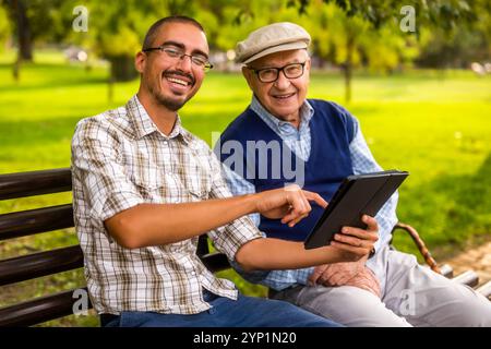 Portrait of happy grandfather and grandson in park. They are spending time together and using digital tablet. Stock Photo