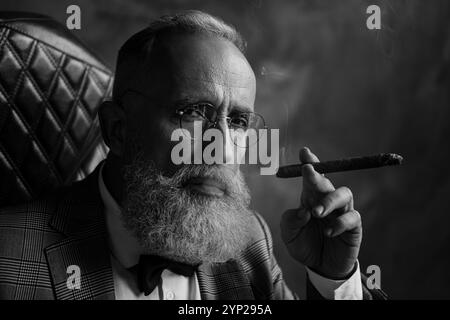 Close up portrait of harsh, brutal, old shareholder in glasses, holding cigar, looking at camera with serious expression, sitting on chair over gray Stock Photo