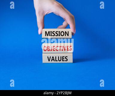 Mission Objectives Values symbol. Concept words Mission Objectives Values on wooden blocks. Beautiful blue background. Businessman hand. Business and Stock Photo