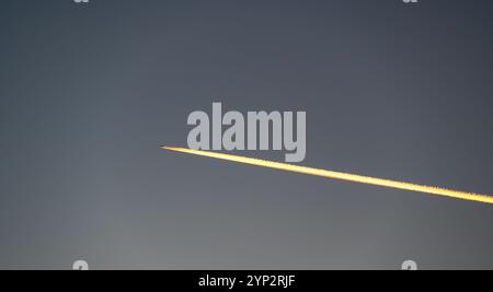 A stunning sunset view of a flying airplane, captured from the ground, with fiery condensation trails glowing in the warm light of the setting sun Stock Photo