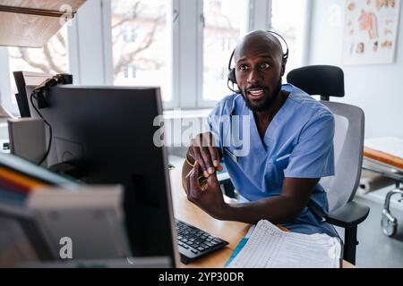Male medical professional wearing headset and assisting patient through video call while sitting on chair in clinic Stock Photo