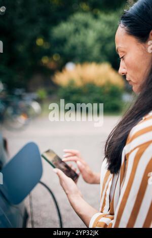 Side view of mature woman using smart phone while charging car in back yard Stock Photo